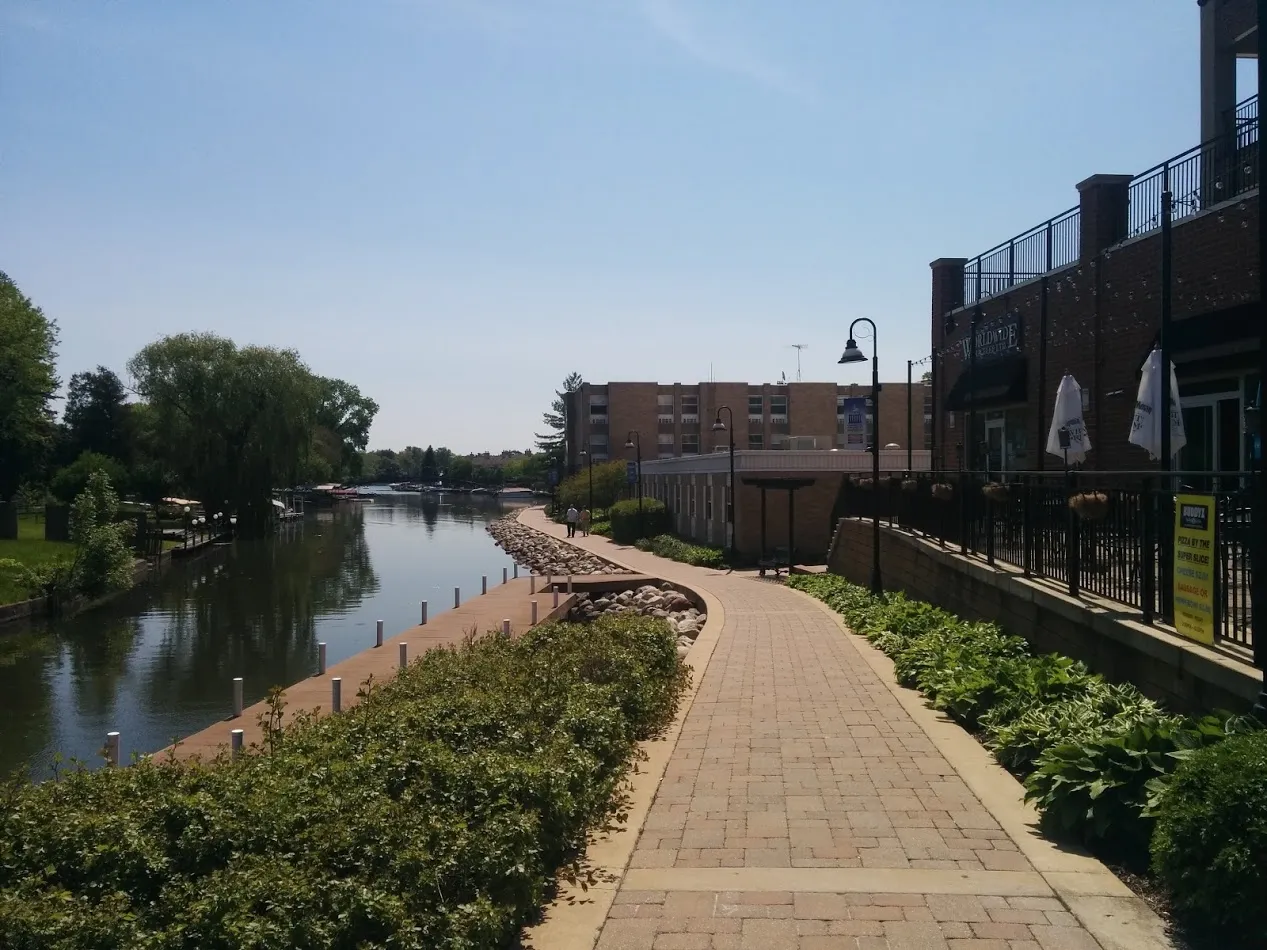 McHenry Riverwalk path beside the water and downtown buildings, looking east from Green Street